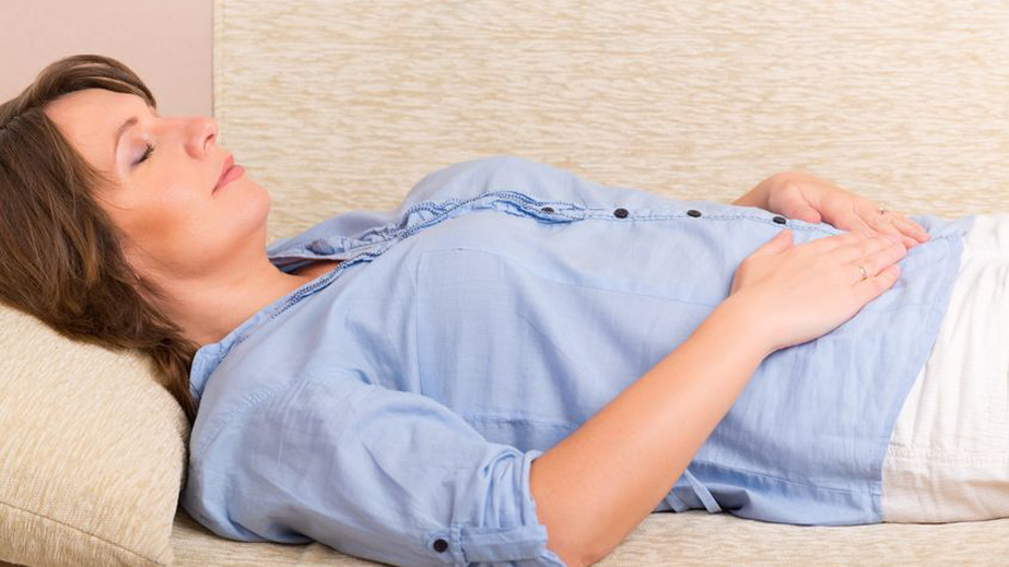 Woman lying on a couch with eyes closed, hands resting on her stomach, wearing a blue shirt and white pants.