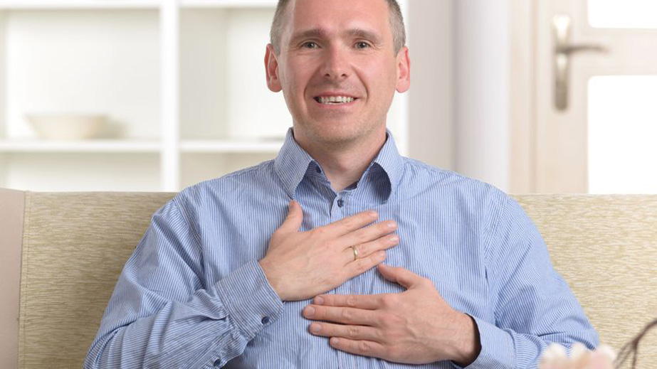 Smiling man sitting indoors places one hand on his chest and another on his stomach, wearing a blue shirt.