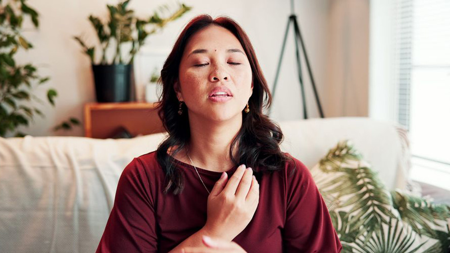 Woman sitting on a couch with eyes closed, touching her chest, appearing to practice deep breathing or meditation.