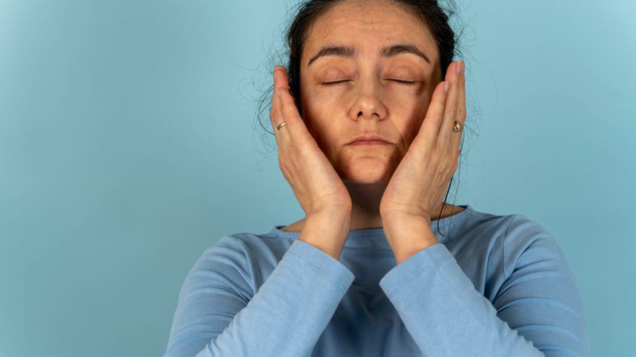 Woman in a blue shirt with closed eyes holds both hands to her face against a plain blue background.