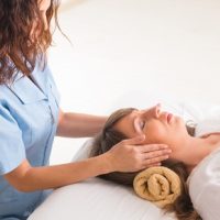 A woman in a white shirt lies on a table with eyes closed while another person massages her head with both hands.