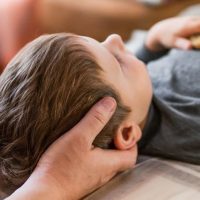 Adult hands cradle a child’s head as the child lies on their back on a bed, facing upward.