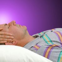 Person lying on a pillow with another person’s hands gently placed on their head, against a purple background.