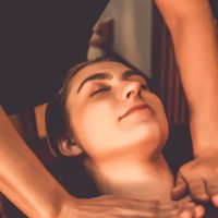 Person with eyes closed receiving a neck and shoulder massage from another individual in a softly lit room.