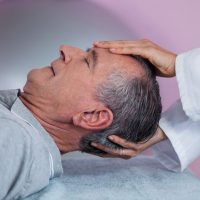 A person in a gray shirt lies on an exam table while a healthcare professional examines their head and neck.